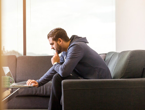 Photo of white male with a trimmed beard sitting on a couch and looking at a computer; for information on how to determine if you bought too much life insurance - from Hunt Insurance, Raleigh.