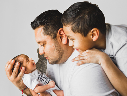 Photo of a Hispanic man holding a newborn baby. His adolescent son is leaning over his shoulder and looking at the baby, too.