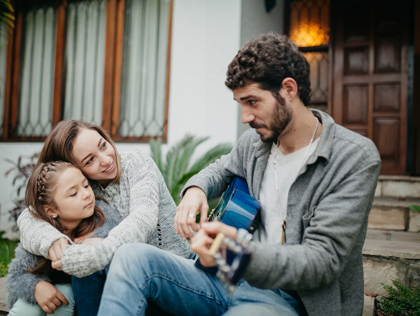 Wife and daughter sitting in front of their house watching Dad play a blue guitar.