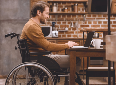 Man in a wheelchair and working on a laptop in the kitchen; for information on who qualitifes for disability benefts