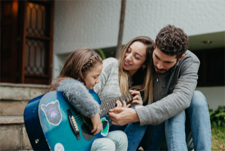 Mother, father, and girl on front steps of their house while dad helps the girl play a guitar; for information on North Carolina whole life insurance from Hunt Insurance.