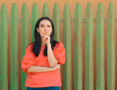 Woman standing in front of a green fence thinking, perhaps about how many life insurance policies she can own